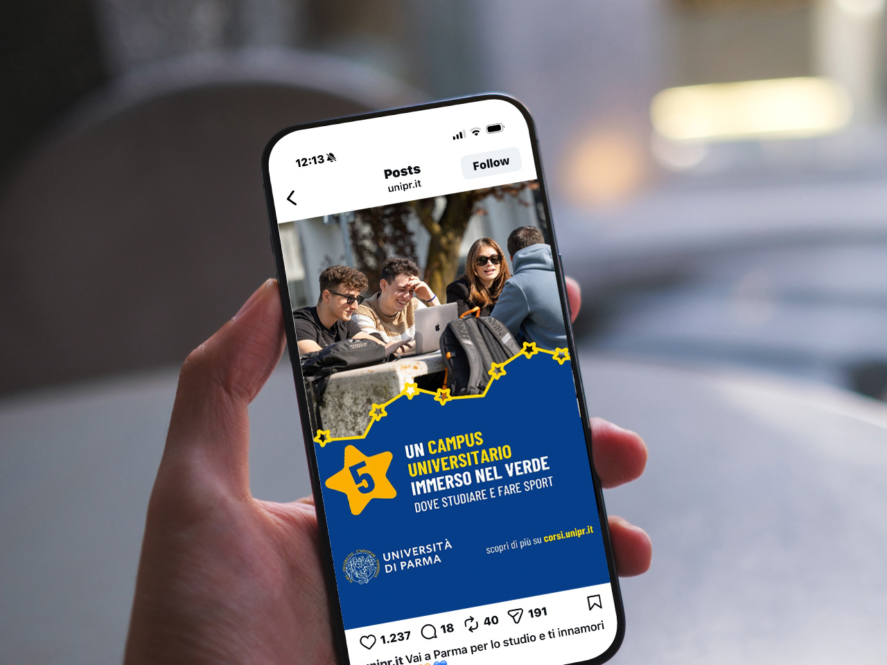 Una mano regge un telefono che mostra un post sui social media dell'università con studenti che studiano all'aperto e un testo in italiano che promuove il campus.