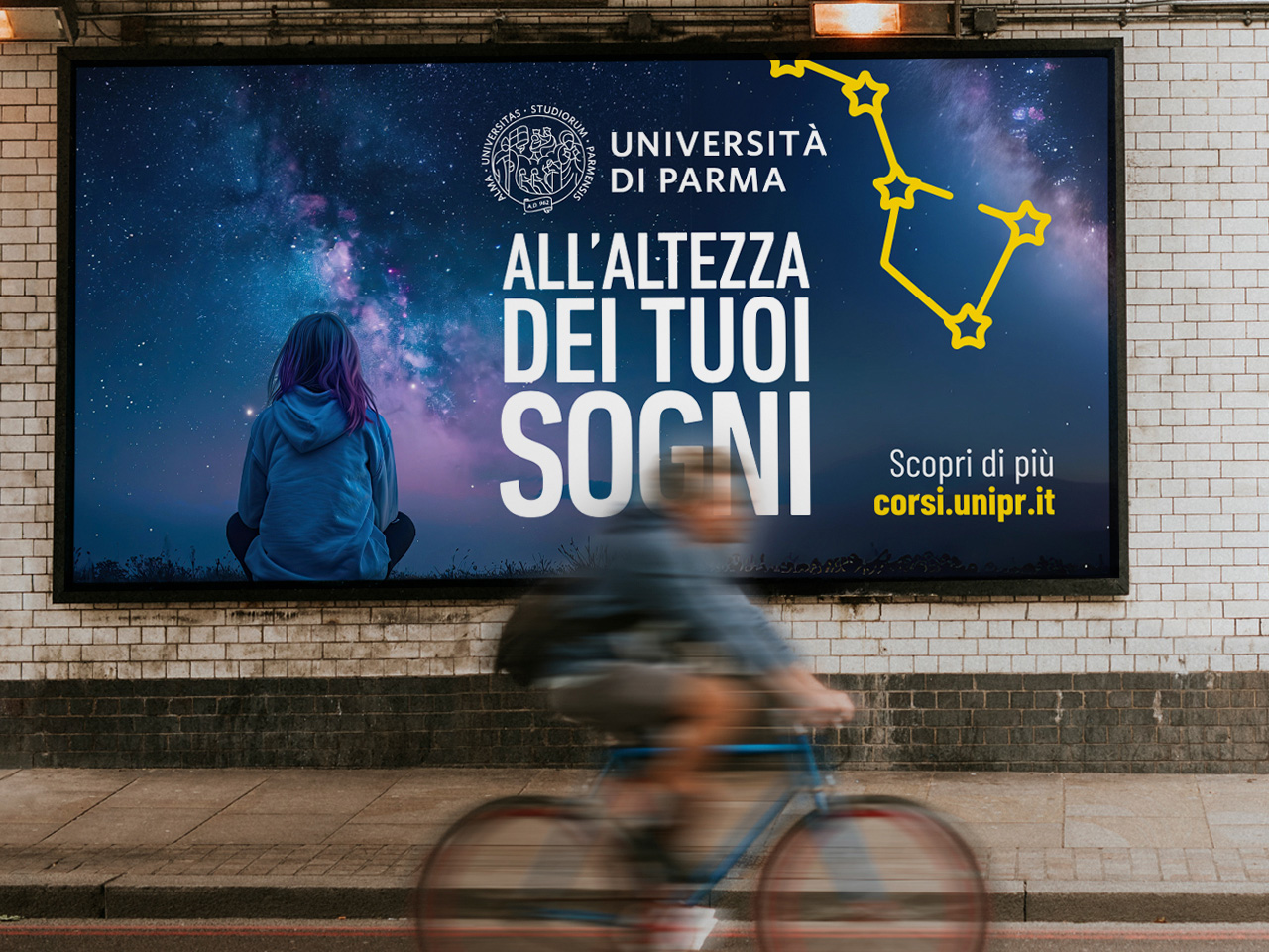 Una persona guarda un cartellone pubblicitario dell'Università di Parma nel cielo stellato; in primo piano passa un ciclista.