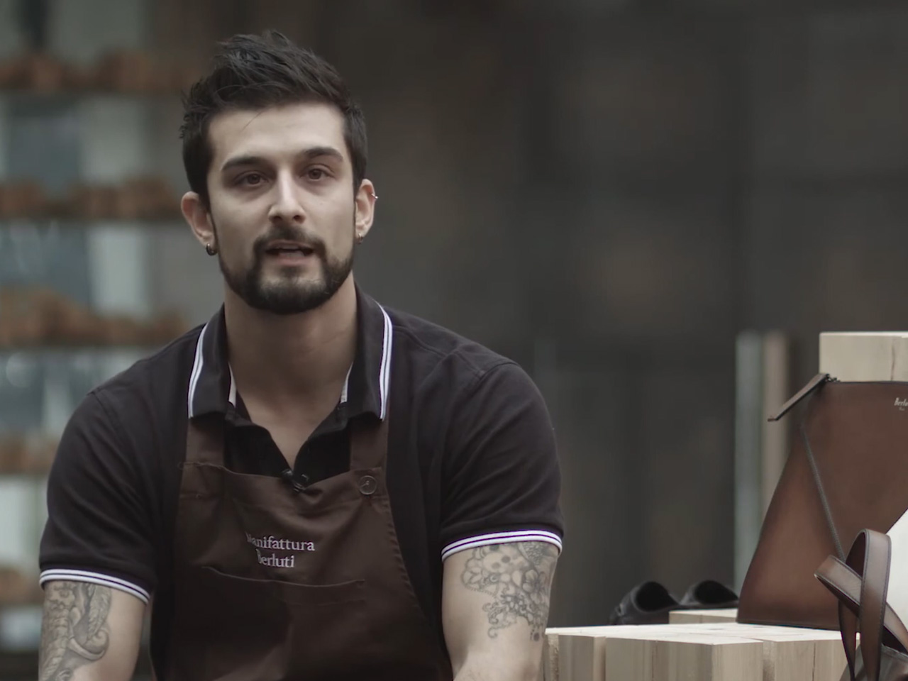 Un uomo con tatuaggi che indossa un grembiule marrone siede accanto a borse di pelle e cassette di legno, guardando leggermente fuori campo.