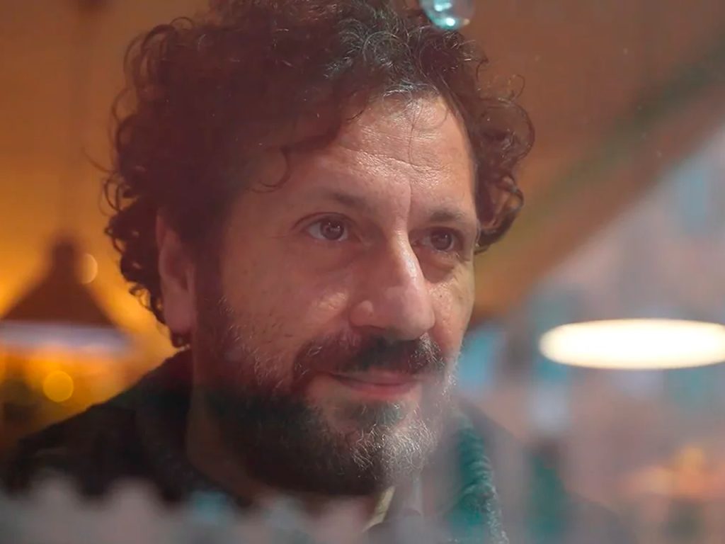 A man with curly hair and beard looks thoughtfully through a window with warm lights in the background.
