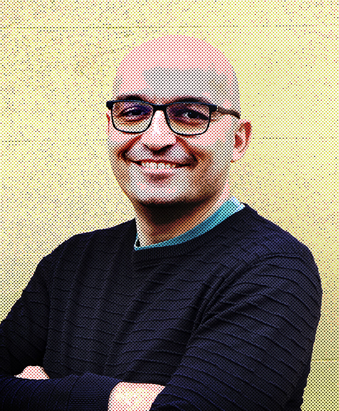 Una persona calva sorridente con occhiali e braccia conserte, che indossa un maglione scuro, su uno sfondo giallo.