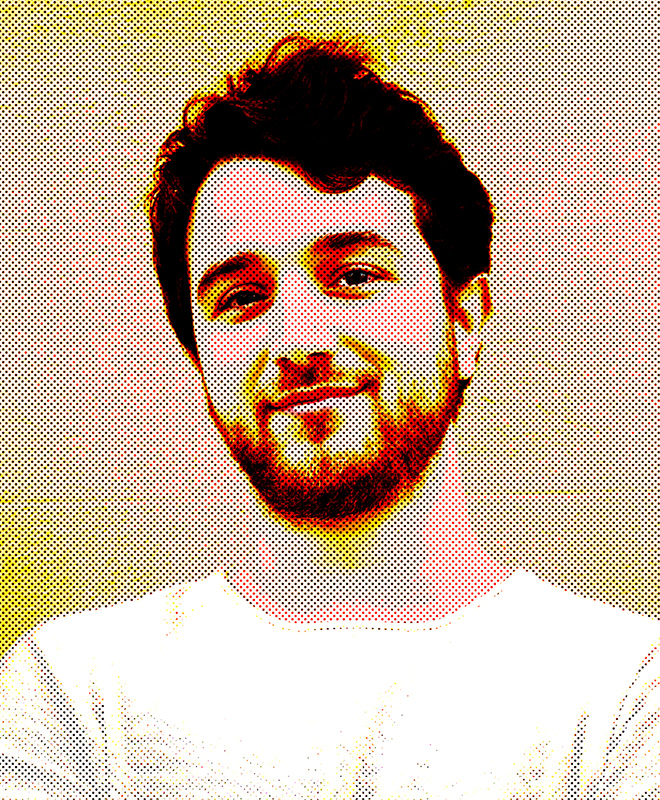 Un uomo barbuto con i capelli scuri sorride dolcemente, indossando una camicia chiara su uno sfondo colorato a puntini.