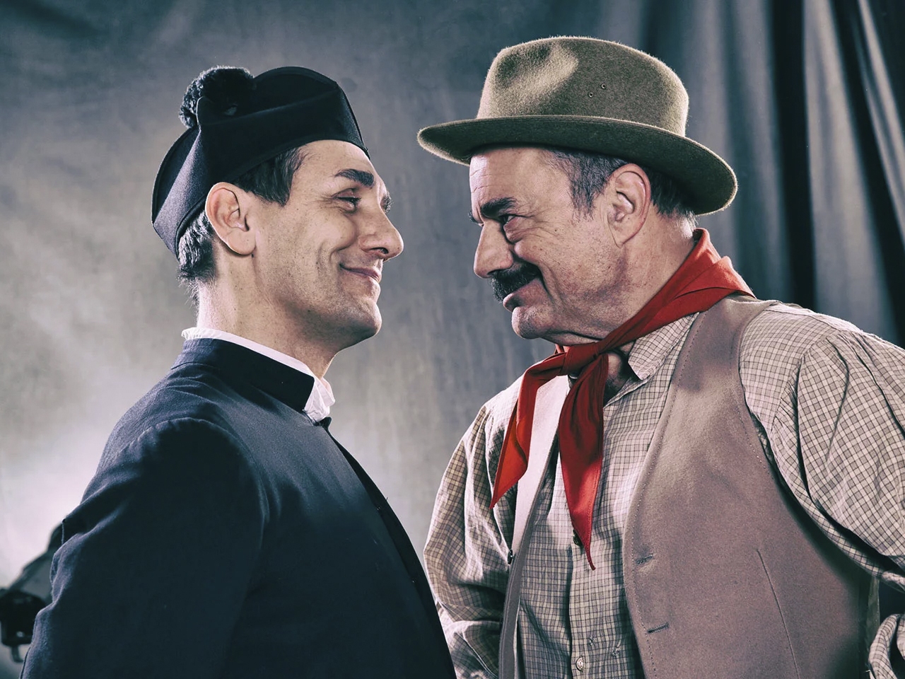 Due uomini in costume d'epoca, uno vestito da prete e l'altro da cowboy, sorridono e si guardano da vicino.