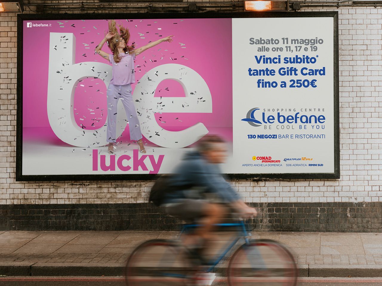 Una donna in rosa si trova accanto a un grande cartello "be lucky"; un ciclista passa accanto a un cartellone pubblicitario di un centro commerciale.
