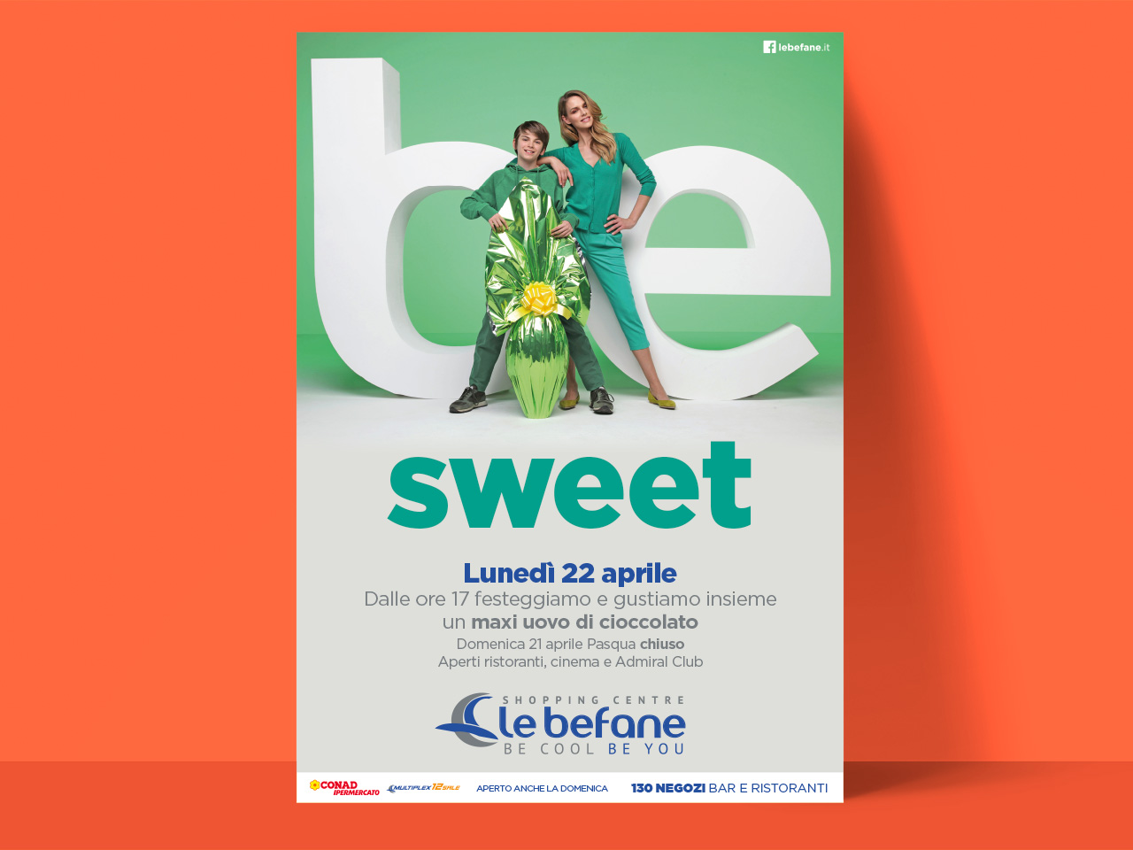 Un ragazzo e una donna sorridenti posano con un uovo di cioccolato gigante davanti a grandi lettere dolci su un poster promozionale.