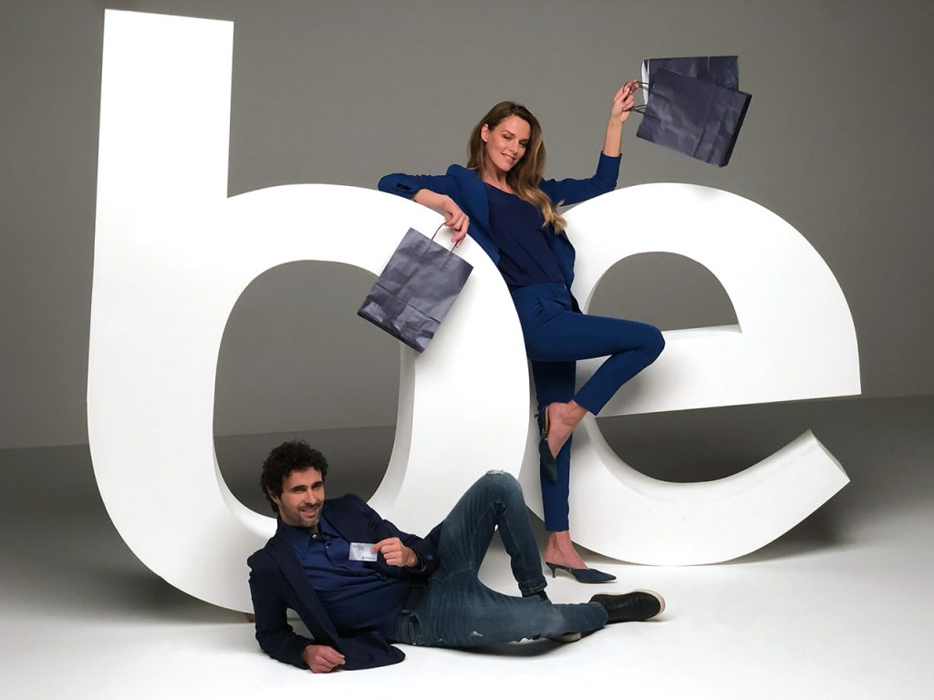 Un uomo e una donna posano con le borse della spesa davanti a grandi lettere bianche scritte su uno sfondo semplice.