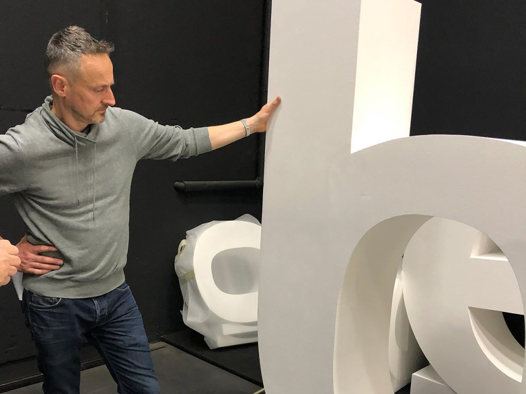 Un uomo con una felpa grigia con cappuccio guarda grandi lettere bianche in 3D in una stanza dalle pareti scure.