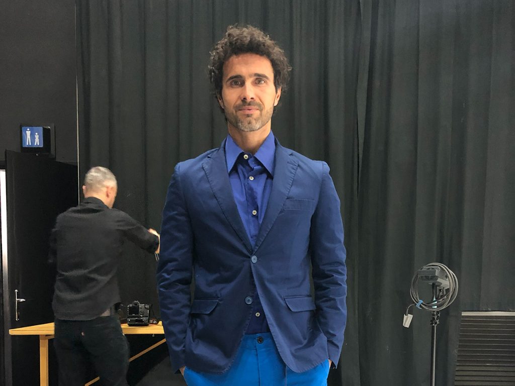 Un uomo in abito blu è in piedi al chiuso; un altro uomo lavora sullo sfondo vicino a un'attrezzatura e a una tenda scura.