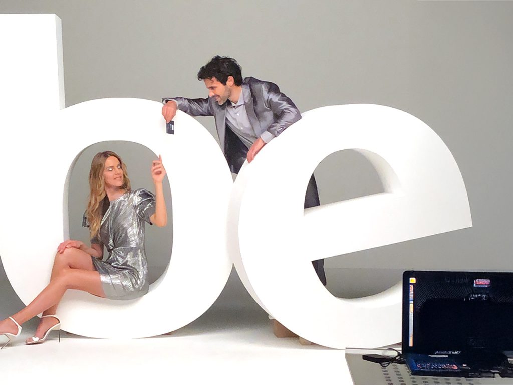 Una donna in abito argentato e un uomo in abito argentato posano con grandi lettere bianche b ed e in uno studio.