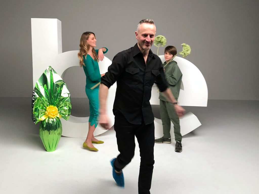 Tre persone posano in uno studio con grandi lettere bianche e una pianta verde lucida; una cammina verso la telecamera.