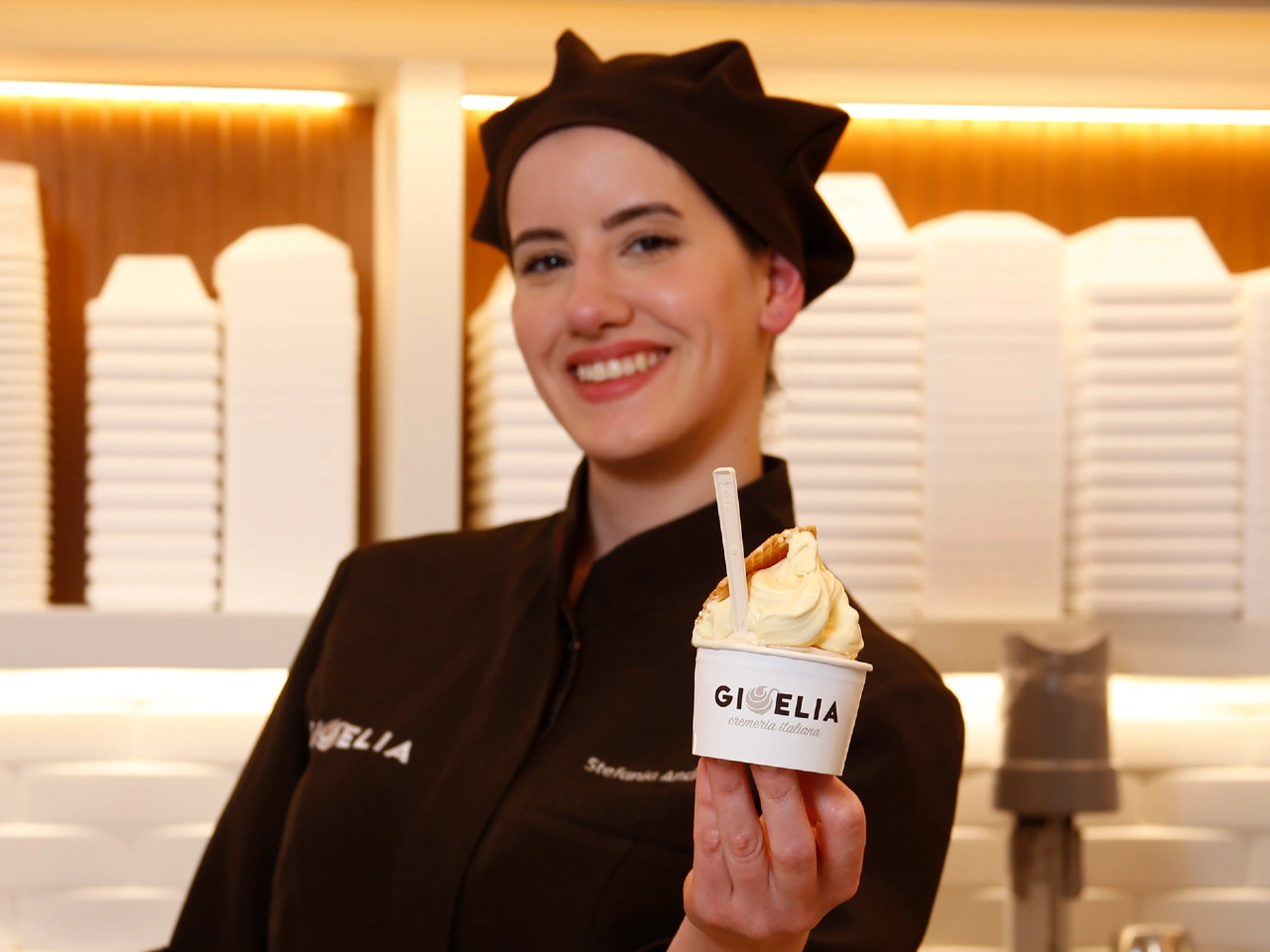 Una cameriera sorridente tiene in mano una coppa di gelato in un negozio, indossando un'uniforme marrone e un cappello.